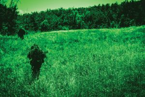 night vision image all green man in camouflage walking through a clearing